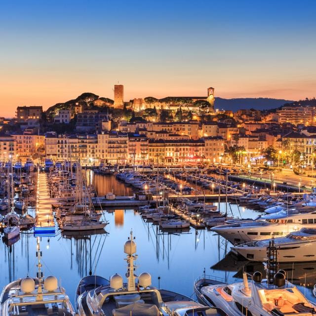 The image showcases a serene view of a marina at dusk, with numerous boats docked along the water. Illuminated buildings line the waterfront, reflecting soft lights in the calm waters. A historic fortress perched on a hill is visible in the background, adding depth to the picturesque scene.