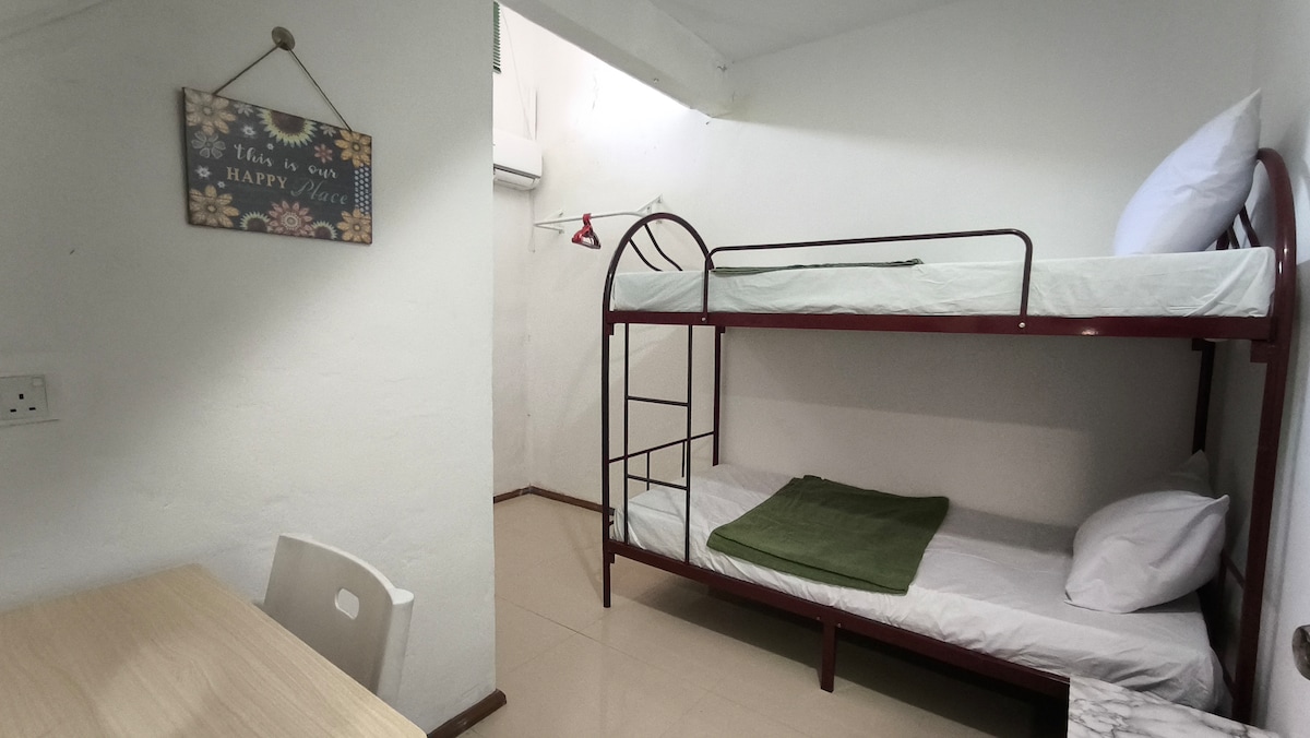 A room features a double-decker bed with a single mattress on the lower level and a thicker mattress on the upper level. A simple table and a green blanket adorn the lower bed. Minimalist decor is present, with a small sign hanging on the wall nearby.
