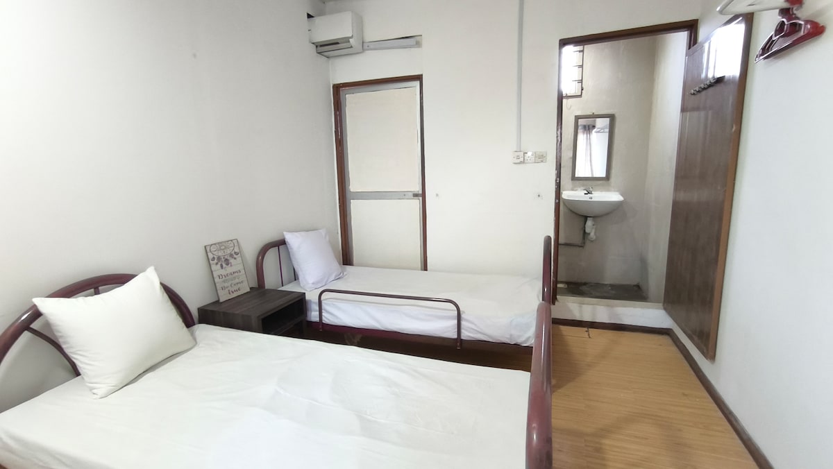 The room is equipped with two single beds positioned parallel to each other. A wooden bedside table is situated next to one bed, providing a practical space for personal items. A wall-mounted mirror and a sink are visible in the background, reflecting a minimalist aesthetic.
