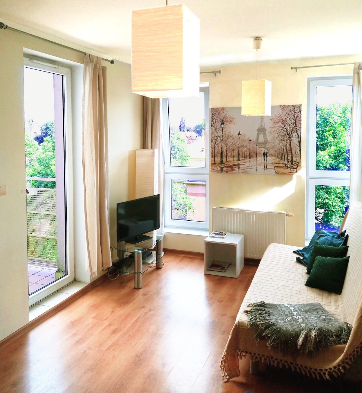 A cozy living area features large windows that invite natural light, complemented by neutral-toned walls. A television is positioned on a sleek stand, while a small table sits beside a green blanket draped over a sofa. A decorative wall art piece adds visual interest.