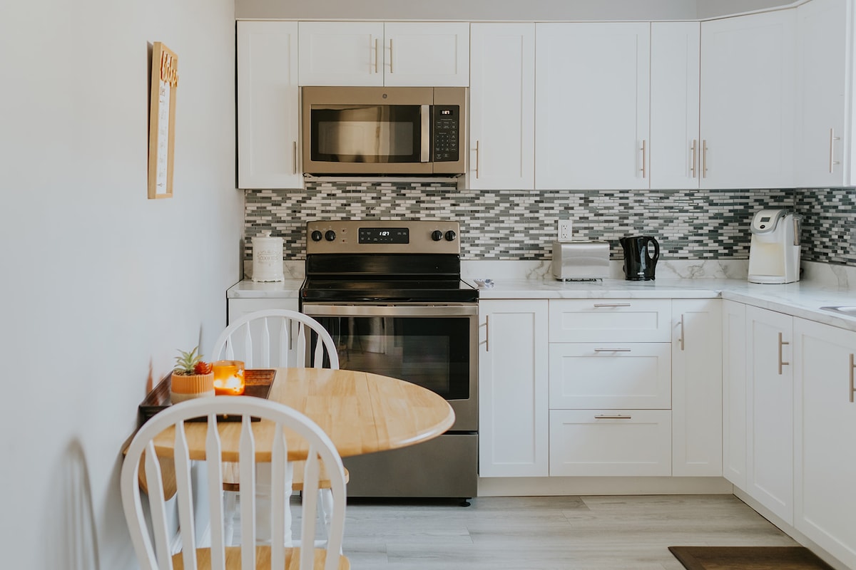 The kitchen features modern white cabinetry and silver appliances, including an oven and microwave. A round wooden dining table with two chairs is positioned against the wall, accompanied by a small succulent plant. Decorative tiles add texture to the backsplash.