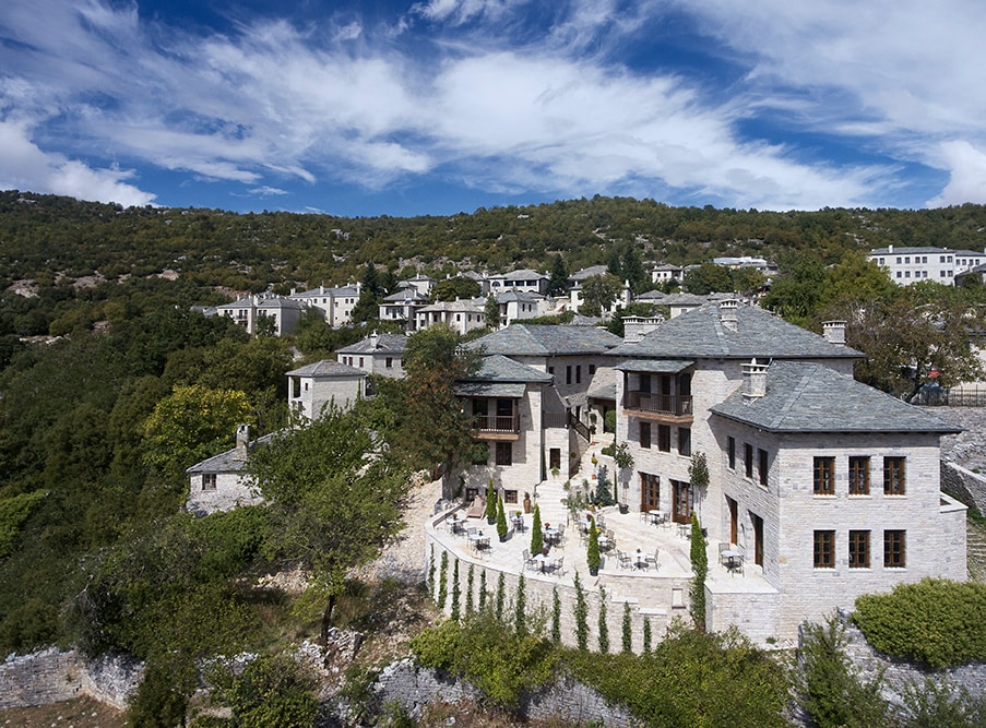A cluster of elegant stone chalets is nestled among lush greenery on a hillside. The architectural design features slate roofs and large terraces surrounded by well-maintained gardens, providing a serene and inviting setting in nature.