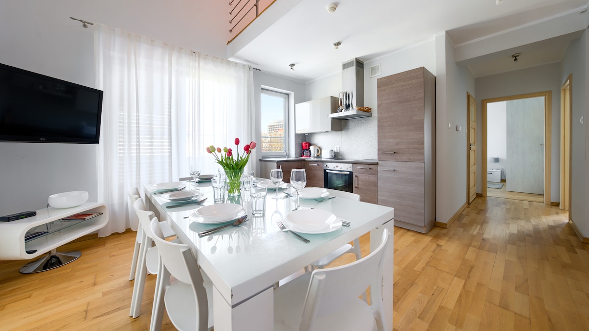 A modern kitchen and dining area is shown, featuring a spacious white dining table set for six. Natural light filters through sheer curtains, illuminating the space. A fully equipped kitchen with dark cabinetry and stainless steel appliances is visible in the background.