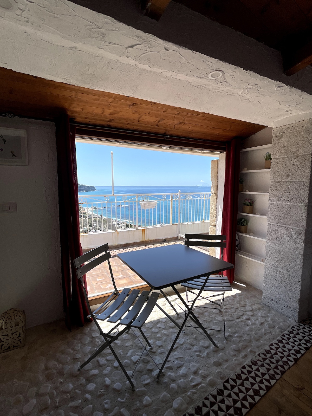 A small dining area features a table with two folding chairs, positioned by a large window. The window frames a clear view of the vast blue sea and coastal landscape, while light pours into the room, enhancing the serene atmosphere.