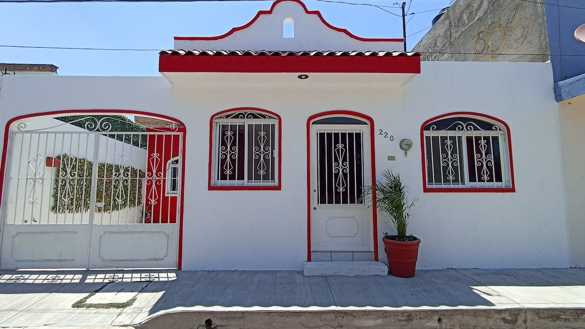 The entrance of a single-story house features a white façade enhanced with red accents. Two decorative windows flank the front door, which is framed by a small potted plant. A secure gate is visible beside the entrance, contributing to a welcoming appearance.
