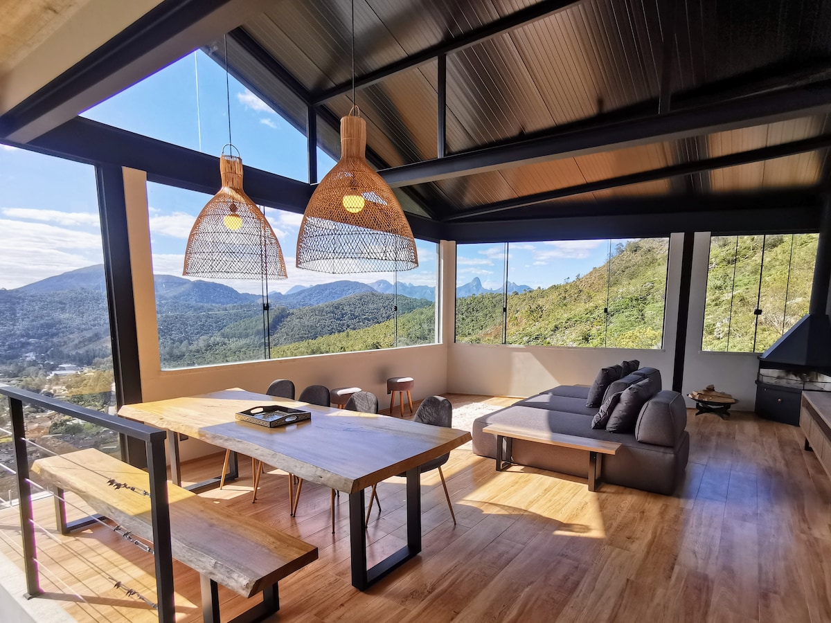 A modern living area features large windows offering expansive views of the surrounding mountains. A wooden dining table is centered in the space, accompanied by simple seating. Two pendant lights hang overhead, while a cozy seating arrangement is positioned near the glass and stone-clad fireplace.