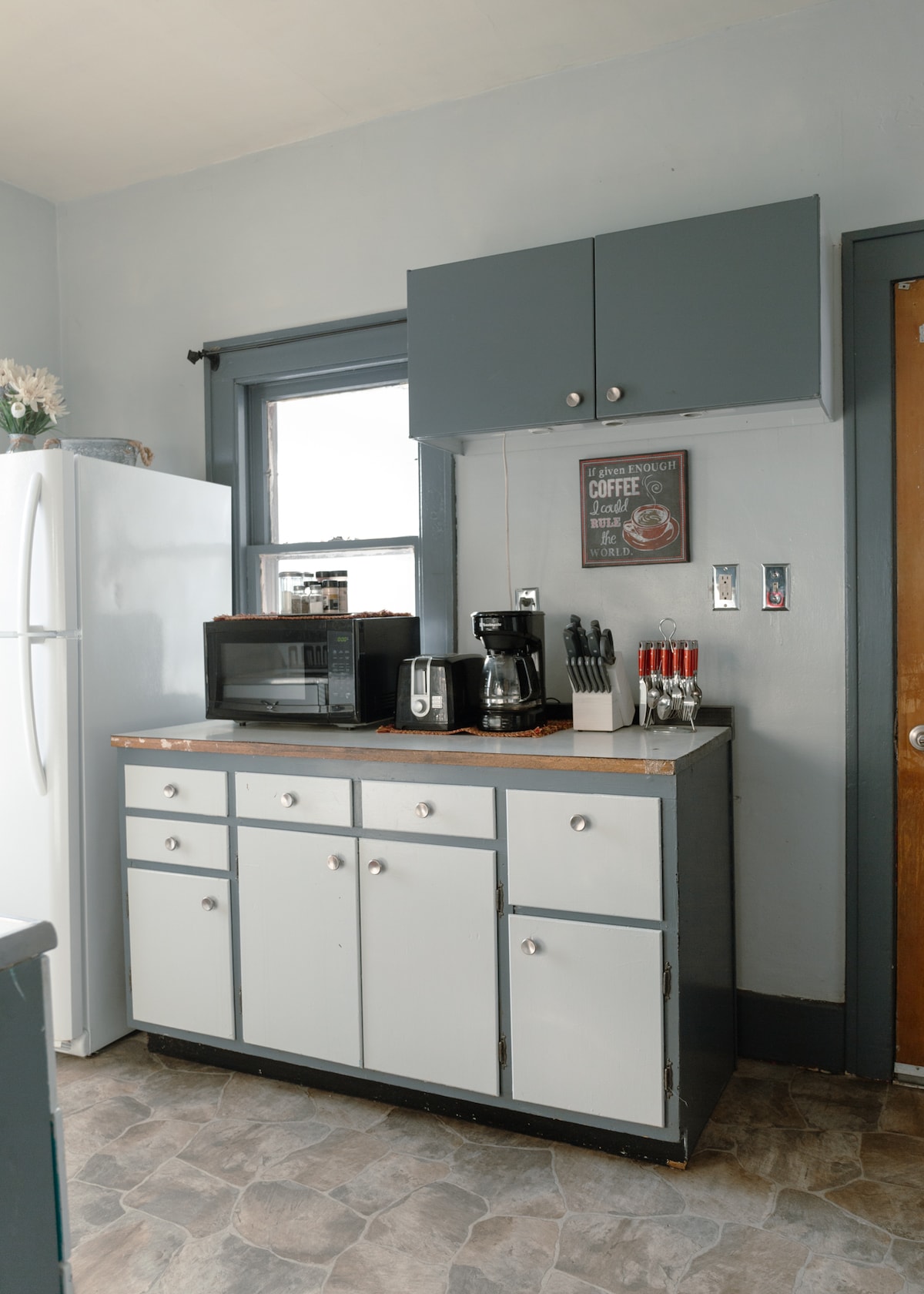 The kitchen showcases a simple design with gray and white cabinetry. A countertop features a coffee maker, microwave, and various utensils. A refrigerator stands to the side, while natural light streams in through a nearby window, highlighting the functional layout.
