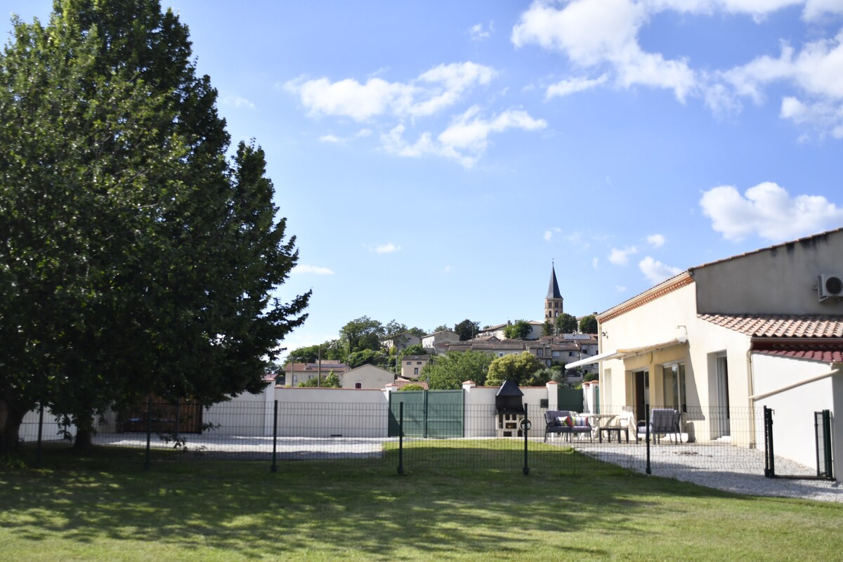 An outdoor area is visible, featuring a lush green lawn enclosed by a fence. A barbecue unit is positioned to the right, while a dining set is arranged nearby. In the background, a quaint church steeple rises above residential buildings under a bright blue sky with scattered clouds.
