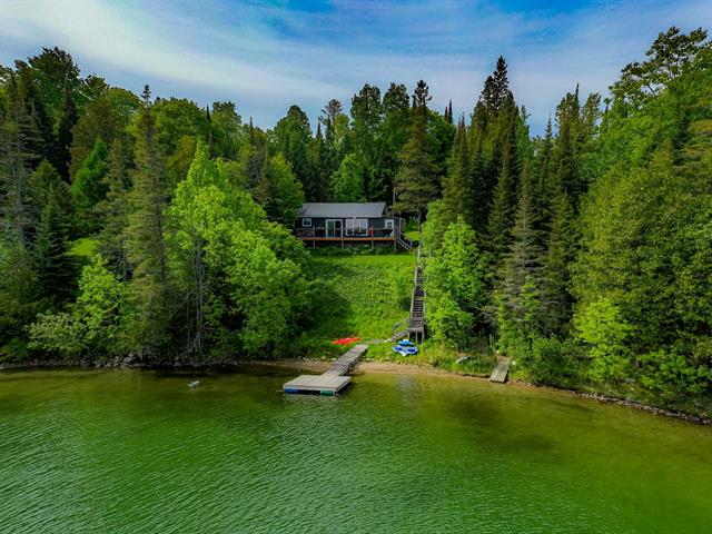 The cottage is set against a backdrop of lush greenery, overlooking the calm waters of Roddick Lake. A private dock extends into the water, bordered by a sandy shoreline. The property features a gradual slope leading from the cottage to the lake, framed by trees.