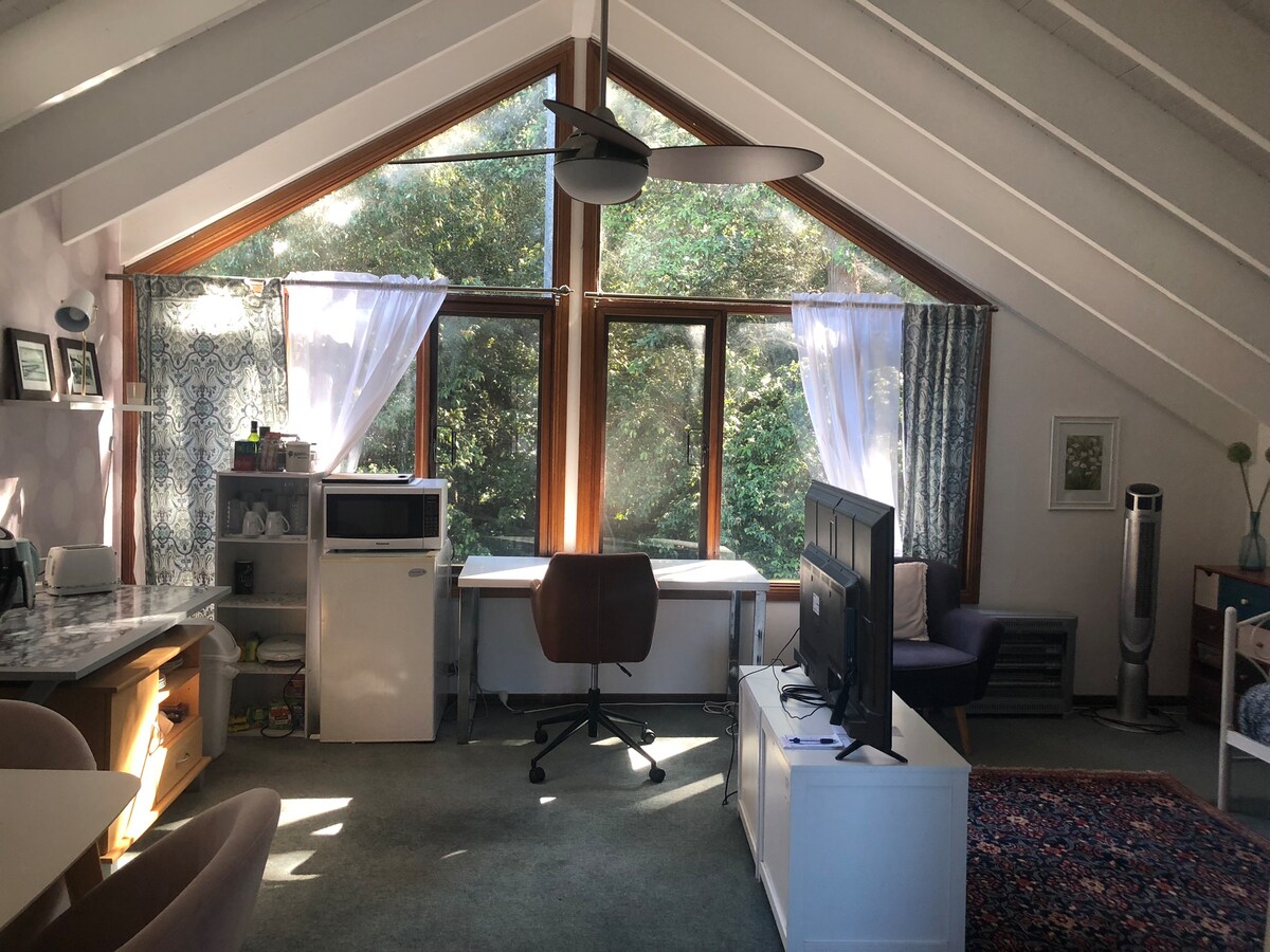 A spacious living area features large windows allowing natural light to fill the room, framed by sheer curtains. A TV is positioned in front of a comfortable couch, with a work desk pairing with a chair. The kitchenette includes a fridge and countertop for meal prep.