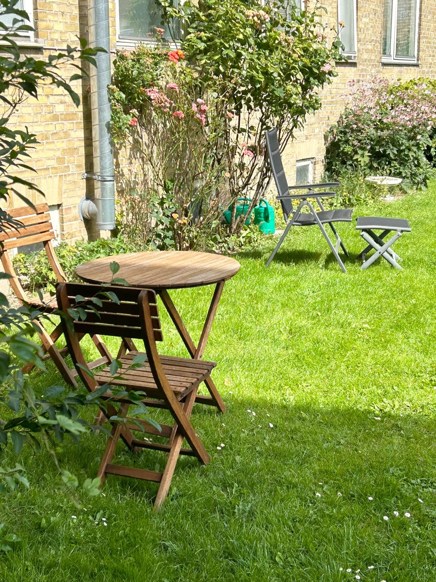 A grassy outdoor area is presented, featuring a small wooden table surrounded by folding chairs. Lush greenery and blooming flowers provide a natural backdrop, enhancing the serene environment. The wall of the building, with brick texture, adds to the overall setting.