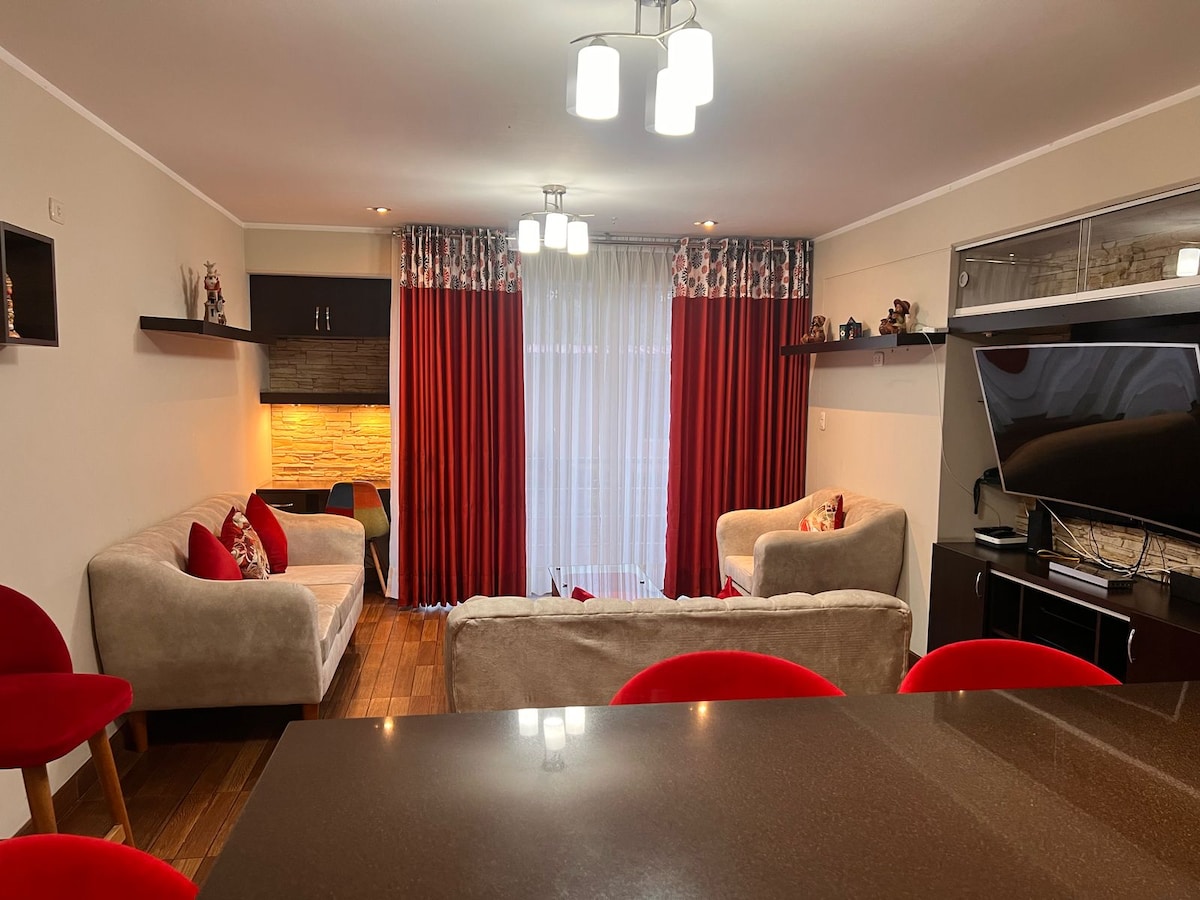 A comfortable living area features two light-colored sofas and a flat-screen TV. Red curtains frame a window allowing natural light. A small built-in shelf displays decorative items, while seating is provided by two round red chairs situated near the wall with soft lighting.