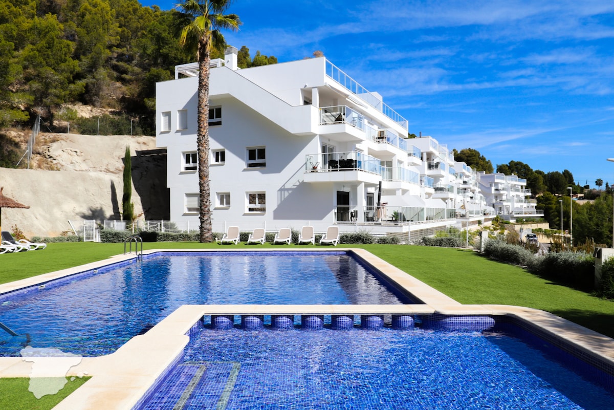 A swimming pool with clear blue water is featured, surrounded by well-maintained green lawns. The modern white buildings of the complex are visible in the background, with balconies that overlook the pool area. Palm trees add a touch of greenery to the scenery.