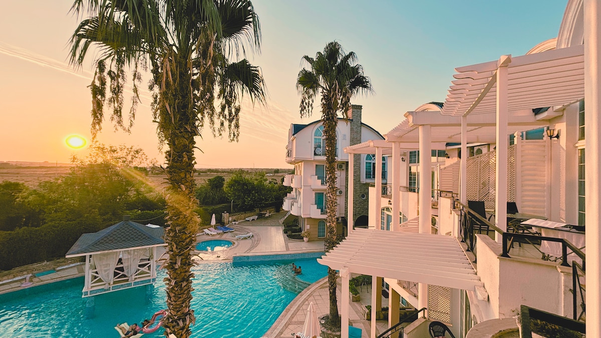 The image captures a tranquil setting by a swimming pool at sunset. Palm trees frame the scene, with two multi-story buildings visible. The pool's inviting blue water reflects the golden hues of the setting sun, creating a peaceful atmosphere. Lounge areas can be seen around the pool.