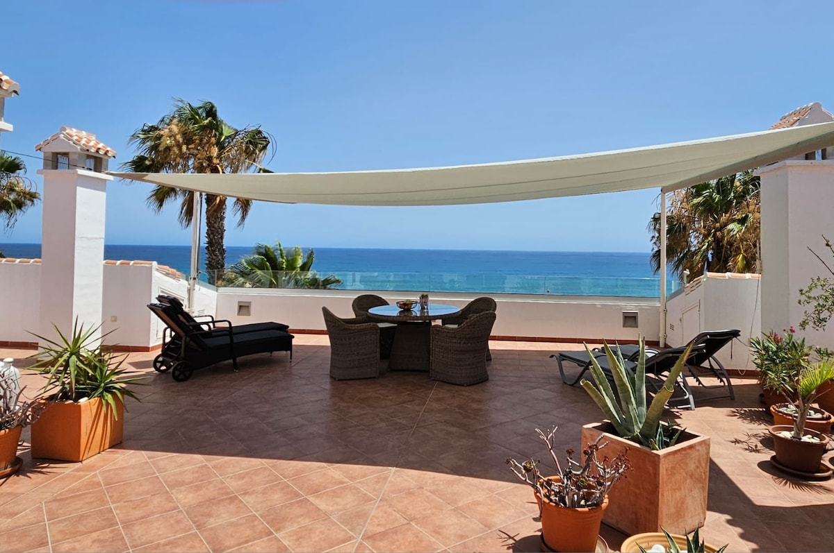 A spacious terrace is featured with a shaded seating area, overlooking a serene ocean view. Potted plants are arranged throughout the space, complementing the coastal surroundings. Comfortable lounge chairs are positioned for relaxation, while a dining table is set for enjoying meals outdoors.