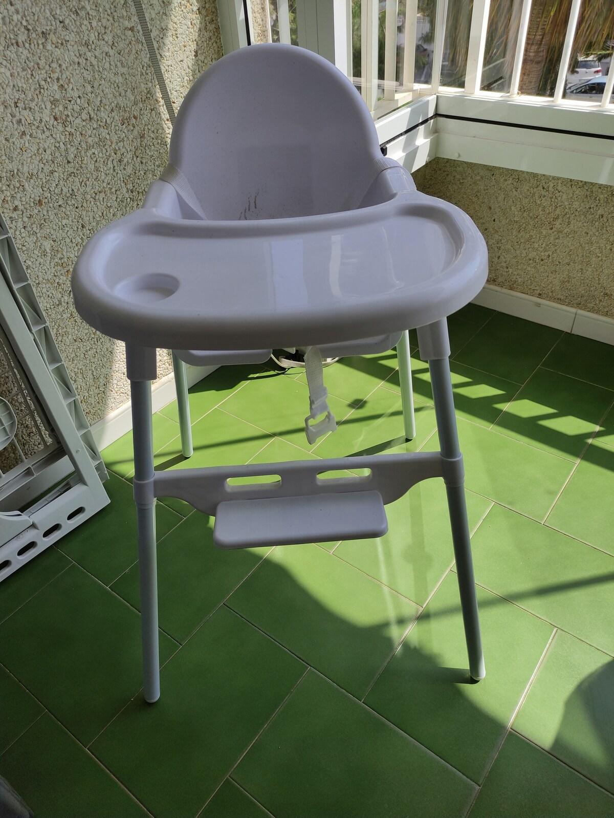 A high chair is positioned on a green tiled floor, featuring a white plastic seat and safety harness. The design includes a small tray for meals and a footrest, contributing to safe dining for young children.
