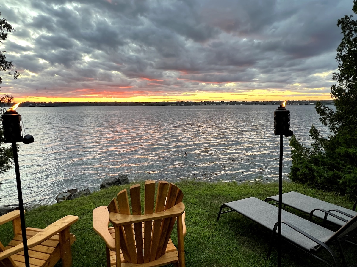 An inviting outdoor space features two wooden Adirondack chairs and two lounge chairs facing the St. Lawrence River. The scene is enhanced by a sunset that casts warm colors across the water, with torches providing ambient lighting against a backdrop of rolling clouds.