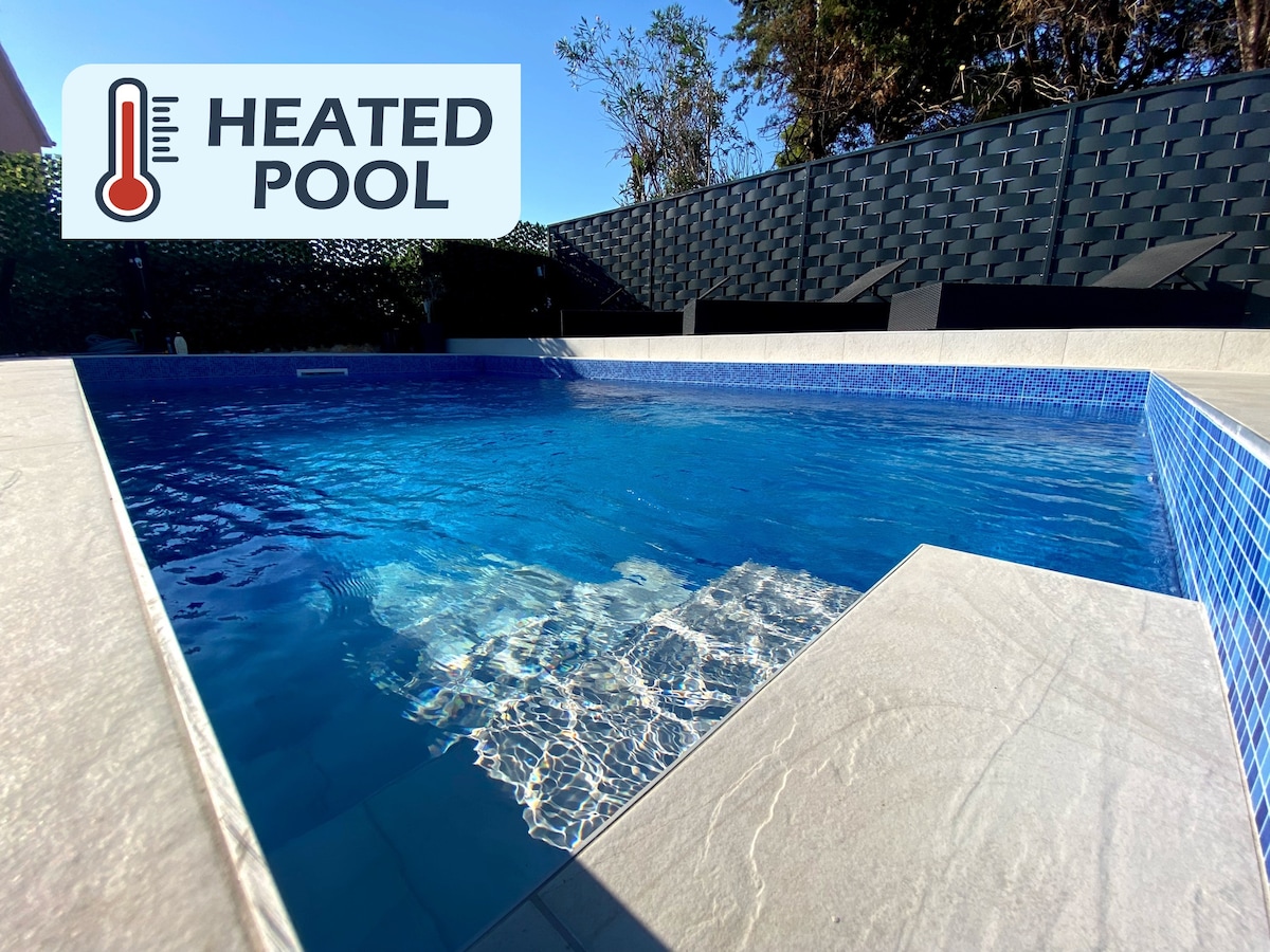 A heated pool is showcased, featuring clear blue water reflecting sunlight. The pool area is surrounded by modern stone tiles, creating a clean and minimalist aesthetic. A textured black wall provides privacy, while lounge chairs are positioned nearby for relaxation.