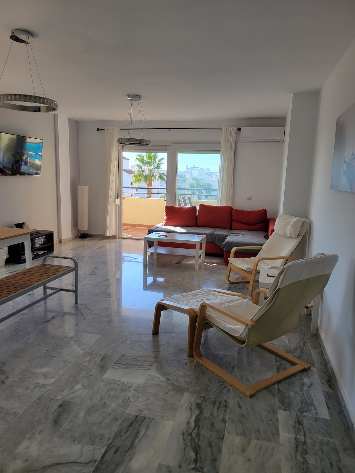 A living area is furnished with a red sofa and two light-colored lounge chairs. A small white table sits in the center. Expansive glass doors provide access to a private balcony, allowing natural light to brighten the marble floor. A television is mounted on the wall.