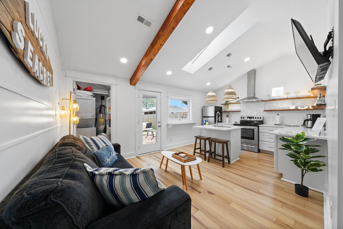 The living area features a comfortable sofa with decorative throw pillows, positioned beside a light wooden coffee table. A sleek kitchen with modern appliances is visible, complemented by vaulted ceilings and abundant natural light from a skylight. Sliding doors lead to a sunny outdoor space.