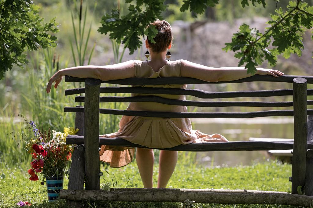 A rustic wooden bench is positioned near a serene body of water, surrounded by lush greenery. A person is seated with arms stretched wide, exuding a sense of tranquility. Colorful flowers are placed beside the bench, adding a natural touch to the peaceful setting.