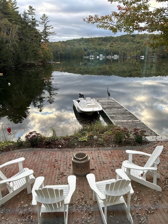 A serene lakeside scene features a wooden deck with four Adirondack chairs arranged around a fire pit. A dock extends into the calm water, with a white boat moored beside it. Reflections of trees and the sky create a tranquil atmosphere.