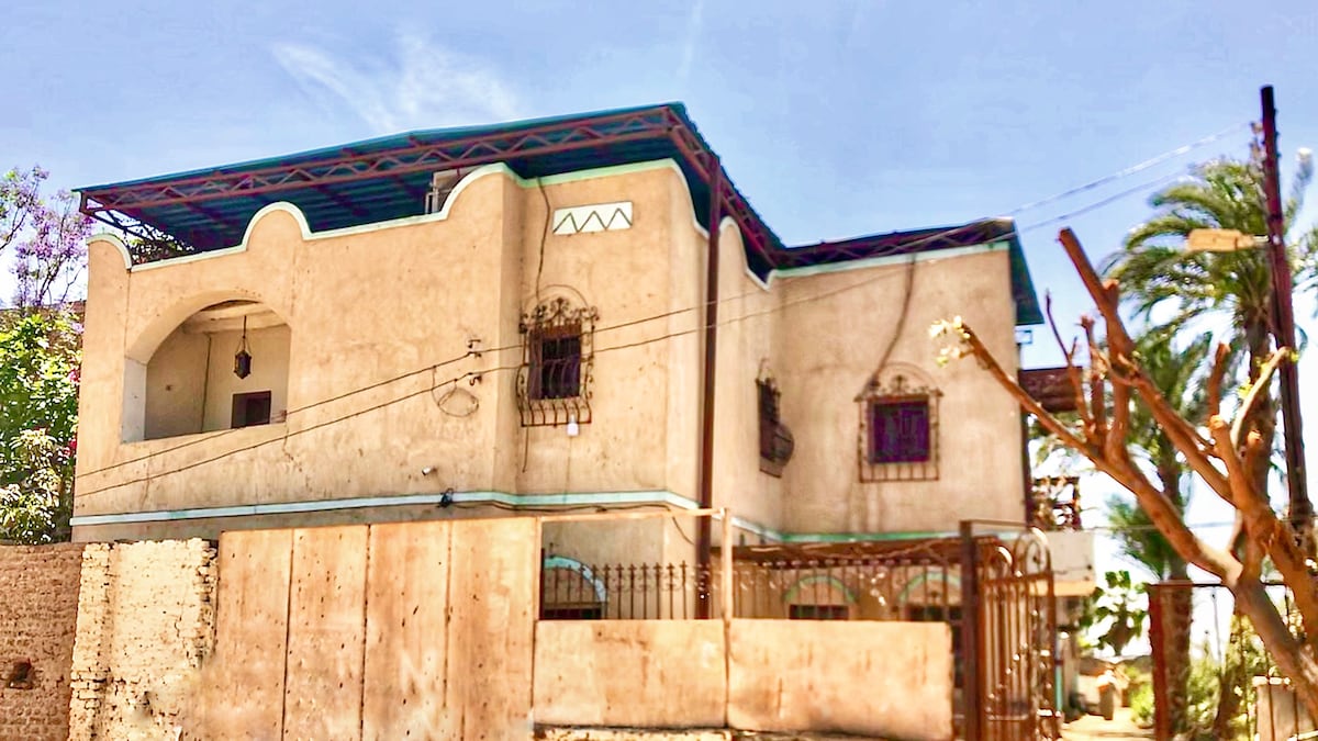 The exterior of a two-story home is showcased, with a textured stucco facade and decorative window grilles. A sloped roof overhangs the upper level, while lush greenery surrounds the property, creating an inviting atmosphere. The entrance is framed with a gated fence for added security.