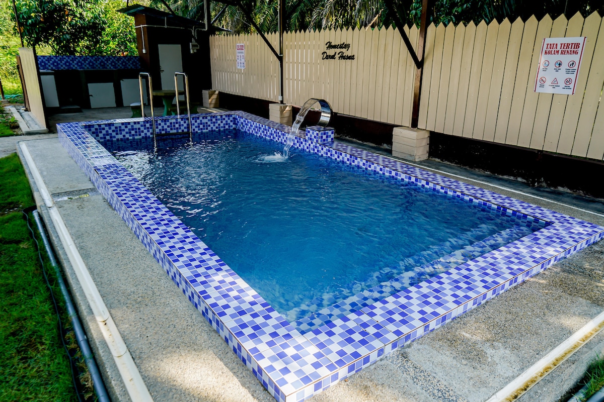 A rectangular pool is seen, featuring blue and white tiled edges with a gentle water fountain creating a tranquil atmosphere. Surrounding the pool are well-maintained concrete surfaces and grassy areas, providing ample space for relaxation.