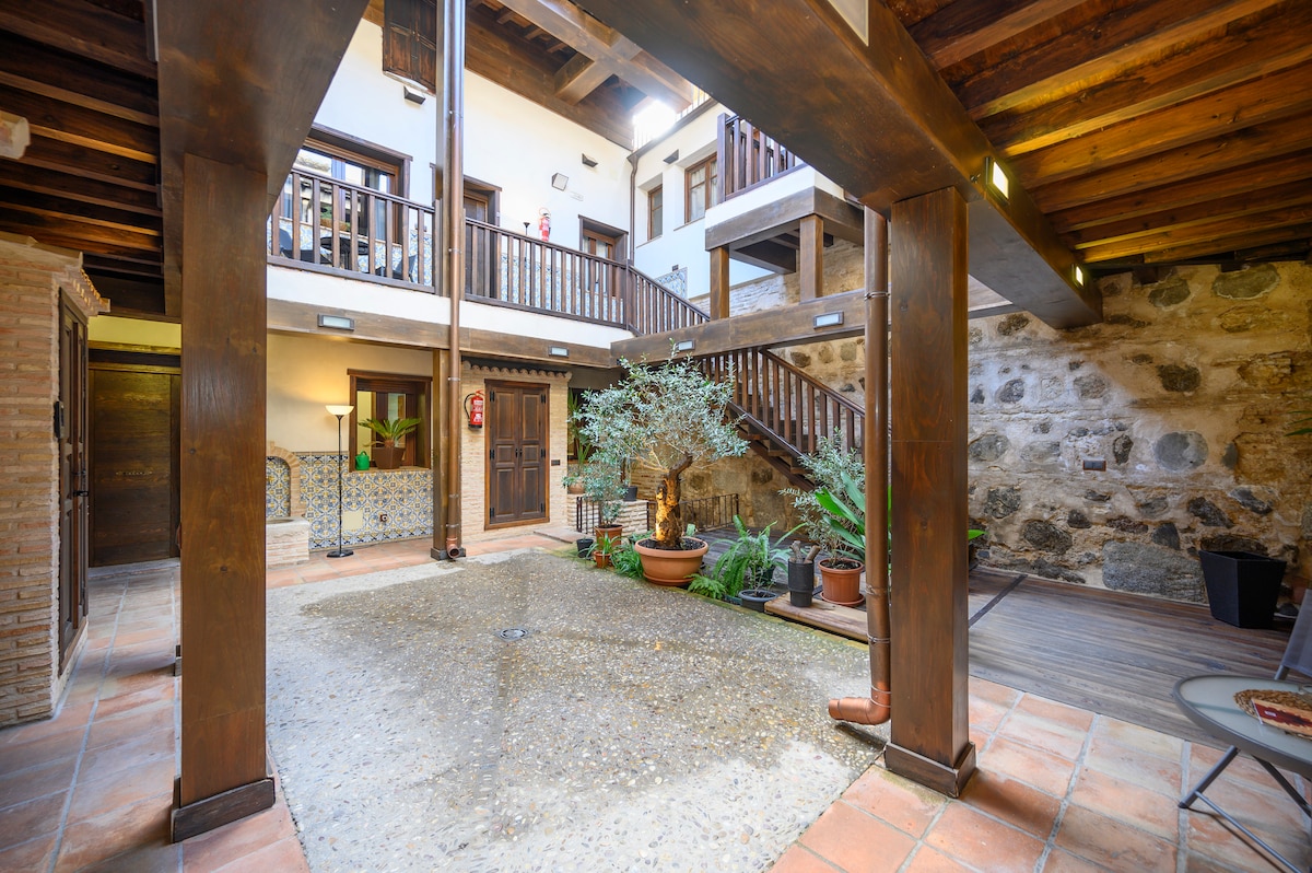 An inviting communal patio area features rustic stone walls and wooden beams. Potted plants and a small tree are strategically placed around a central water feature. Natural light streams in from the upper balconies, enhancing the serene atmosphere of this shared outdoor space.