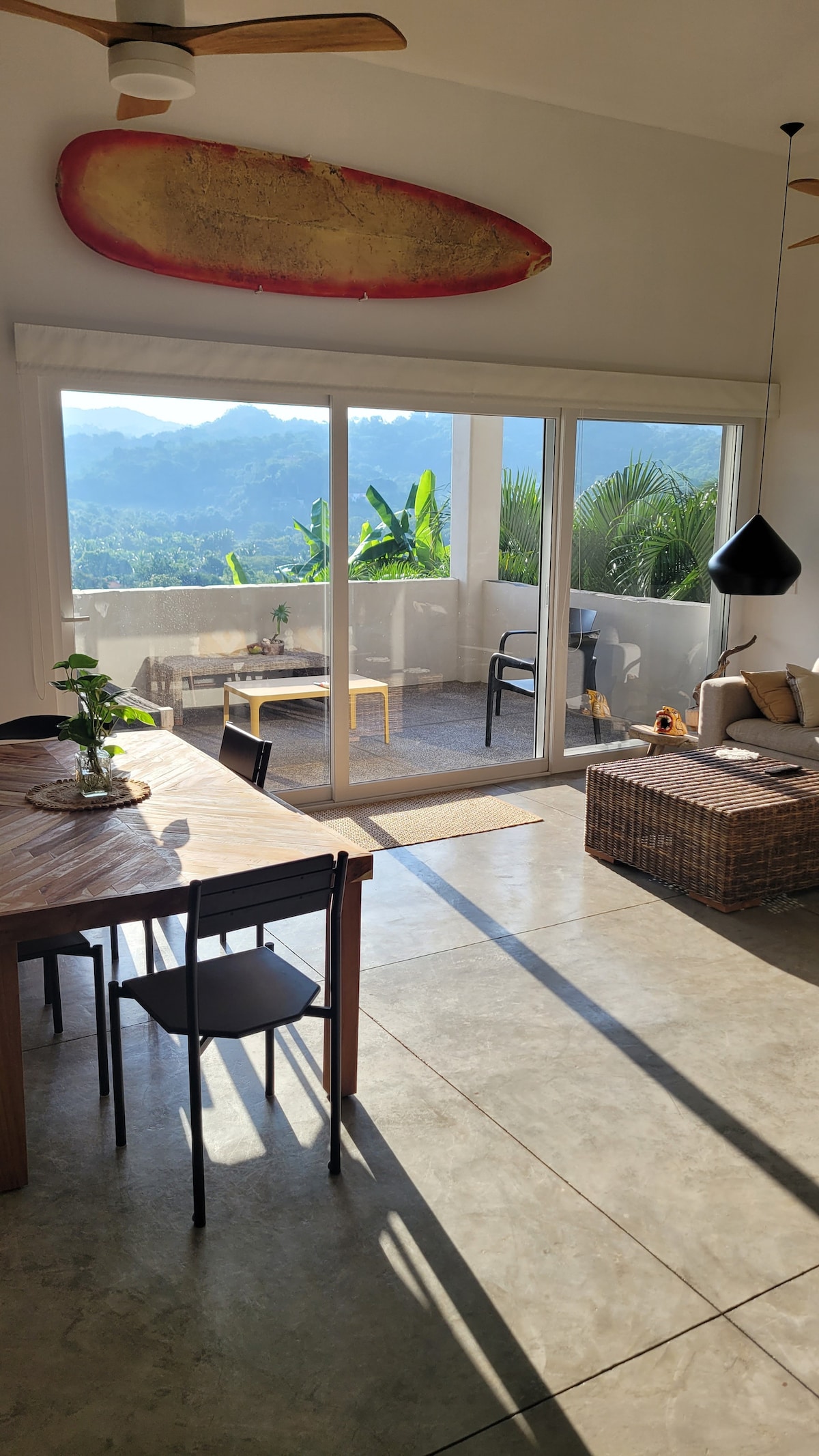 An open living area features a wooden dining table accompanied by black chairs. A surfboard is mounted on the wall, and large glass doors provide access to a terrace showcasing lush mountain views. Natural light fills the space, highlighting the polished concrete floor.