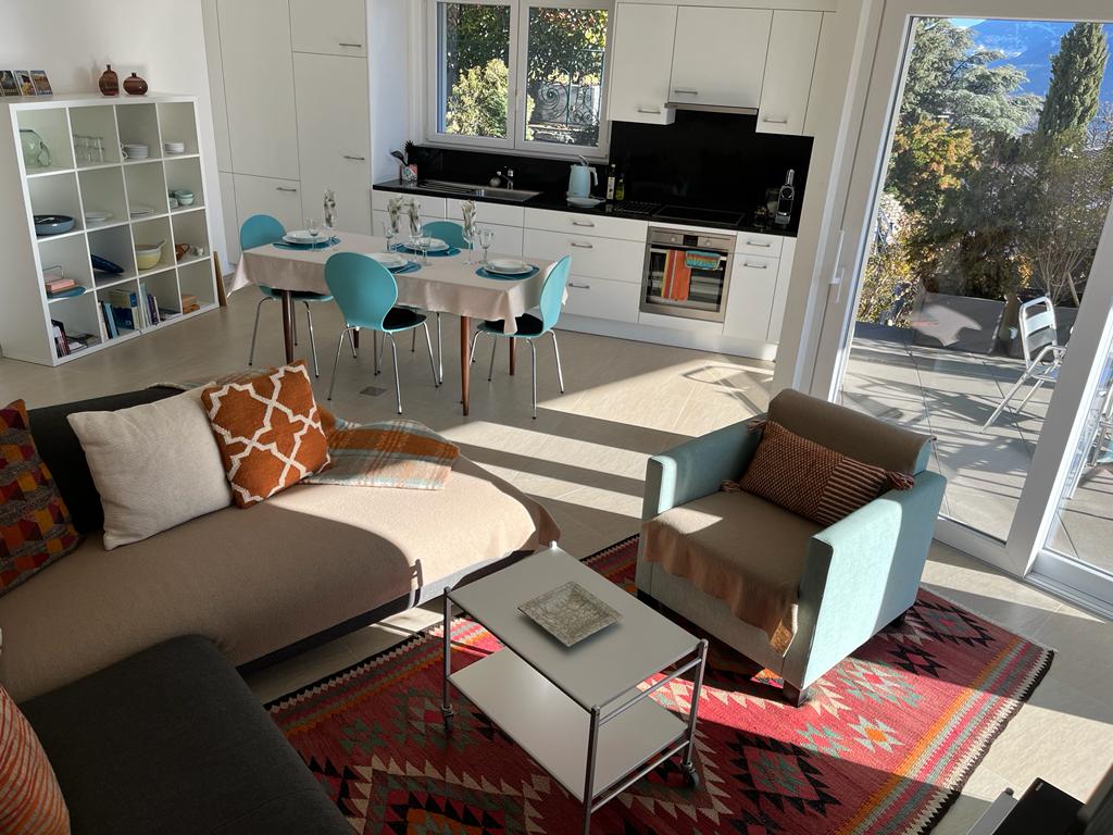 A modern living area showcases a cozy sectional sofa adorned with decorative pillows, set against a vibrant rug. A dining table for six is positioned nearby, complemented by aqua-colored chairs. Large windows allow natural light to fill the space, with an outdoor terrace visible through the glass doors.