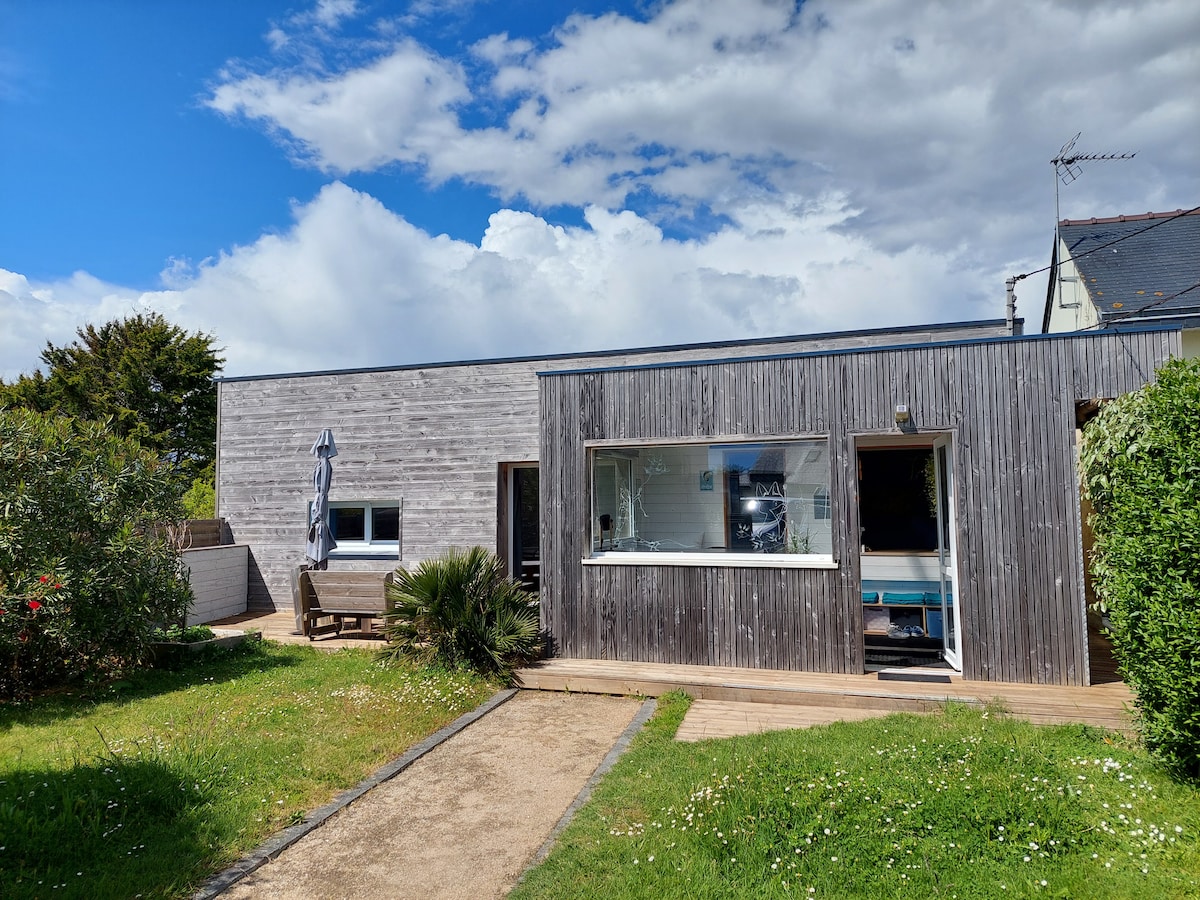 The exterior of a recently renovated house is shown, featuring wooden cladding and large windows. A small patio area with seating faces the garden. Lush greenery surrounds the property, and a pathway leads to the entrance, under a partly cloudy sky.