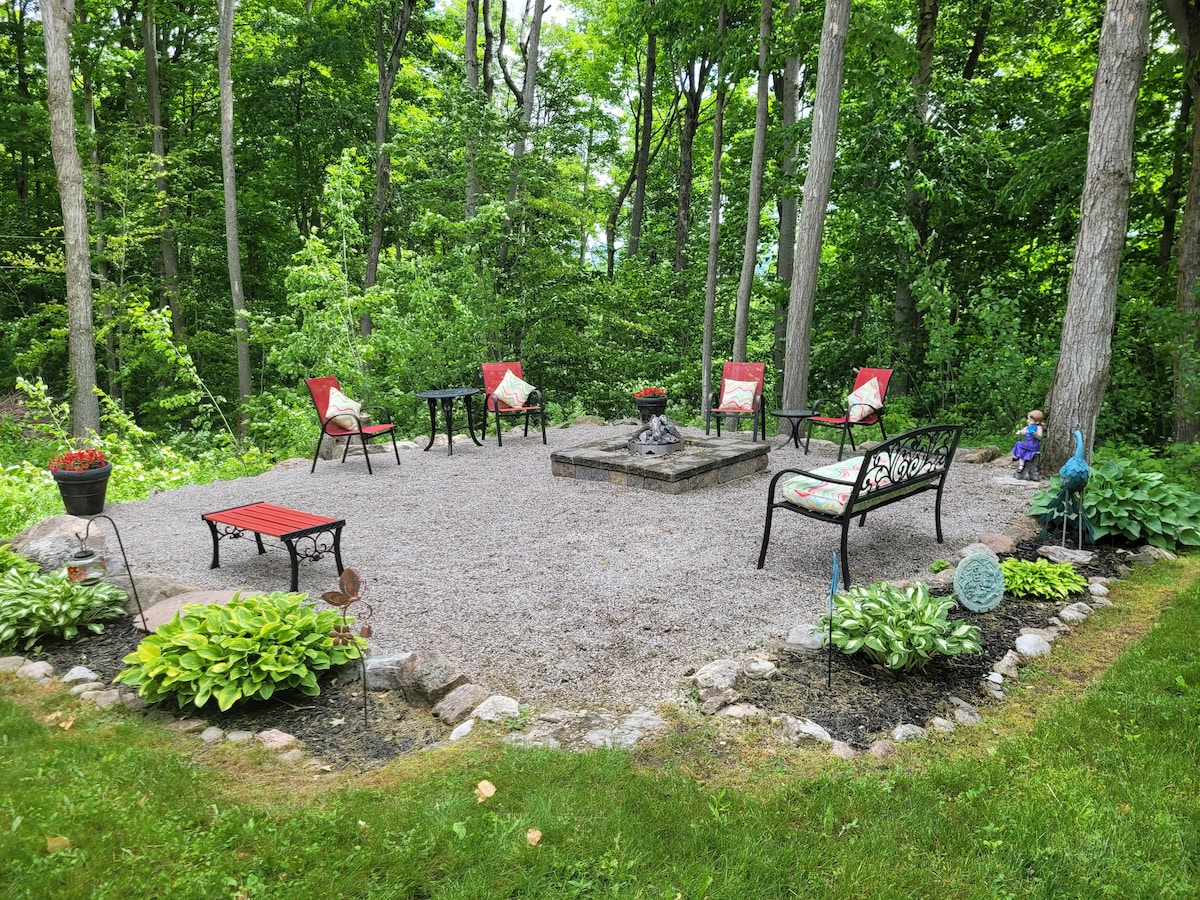 An outdoor seating area is surrounded by lush greenery, featuring a central fire pit surrounded by stone and gravel. Six chairs with red cushions are arranged nearby, complemented by potted plants and decorative stones, creating a serene environment for relaxation.