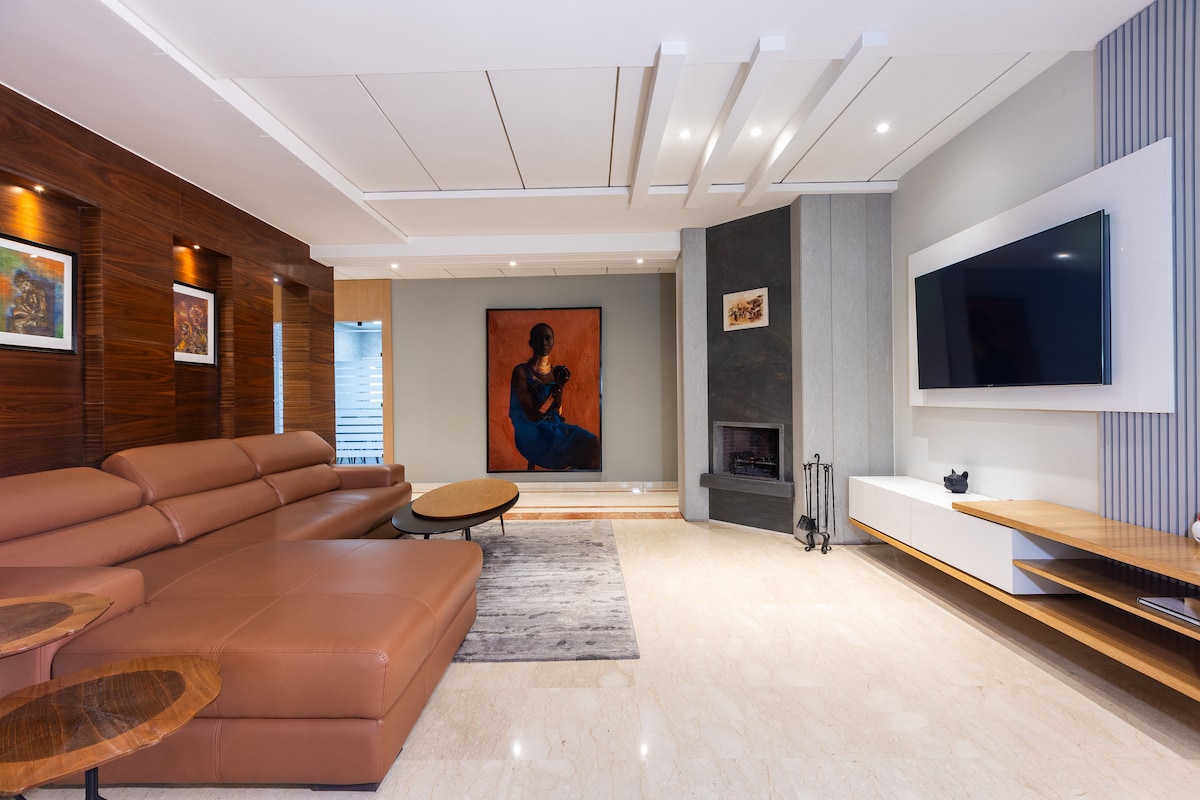 The living area is designed with a large, brown sectional sofa and features a modern coffee table. A piece of artwork is mounted on the wall, and a flat-screen television is positioned above a sleek cabinet. Ambient lighting creates a relaxed atmosphere.