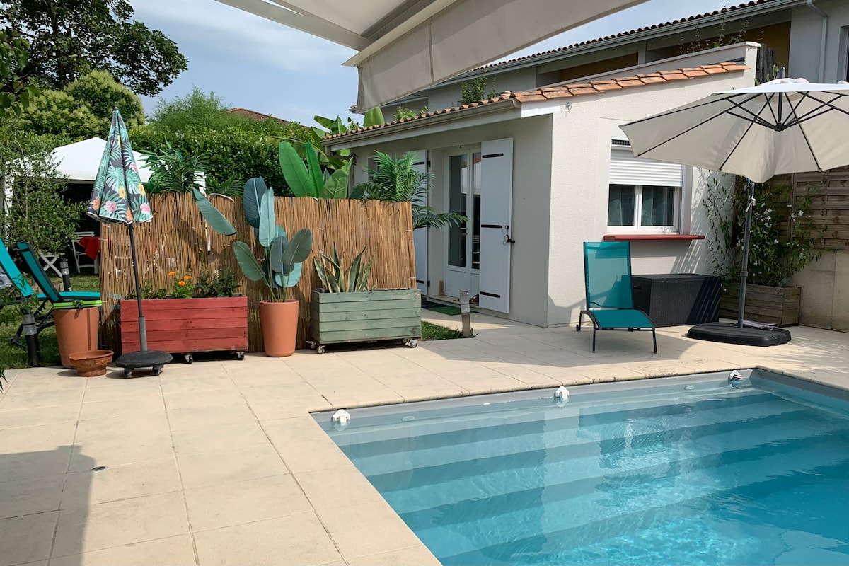 A pool area is bordered by greenery and colorful planters. Comfortable lounge chairs and an umbrella provide shaded seating. A spacious, clear pool reflects the blue sky, while a well-maintained exterior of the studio is visible in the background.