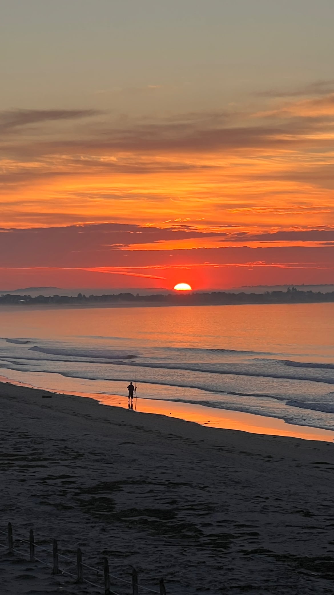 A serene beach scene at sunset, with vibrant orange and pink hues illuminating the sky. A lone figure stands along the wet sand, reflecting the colors of the sunset. Gentle waves lap at the shore, and silhouettes of distant trees can be seen against the horizon.