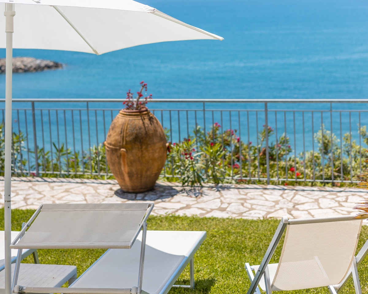 A spacious terrace is presented, featuring sun loungers and a large umbrella offering shade. An ornate clay amphora is situated amidst lush greenery, providing a charming focal point against the backdrop of the serene sea view.