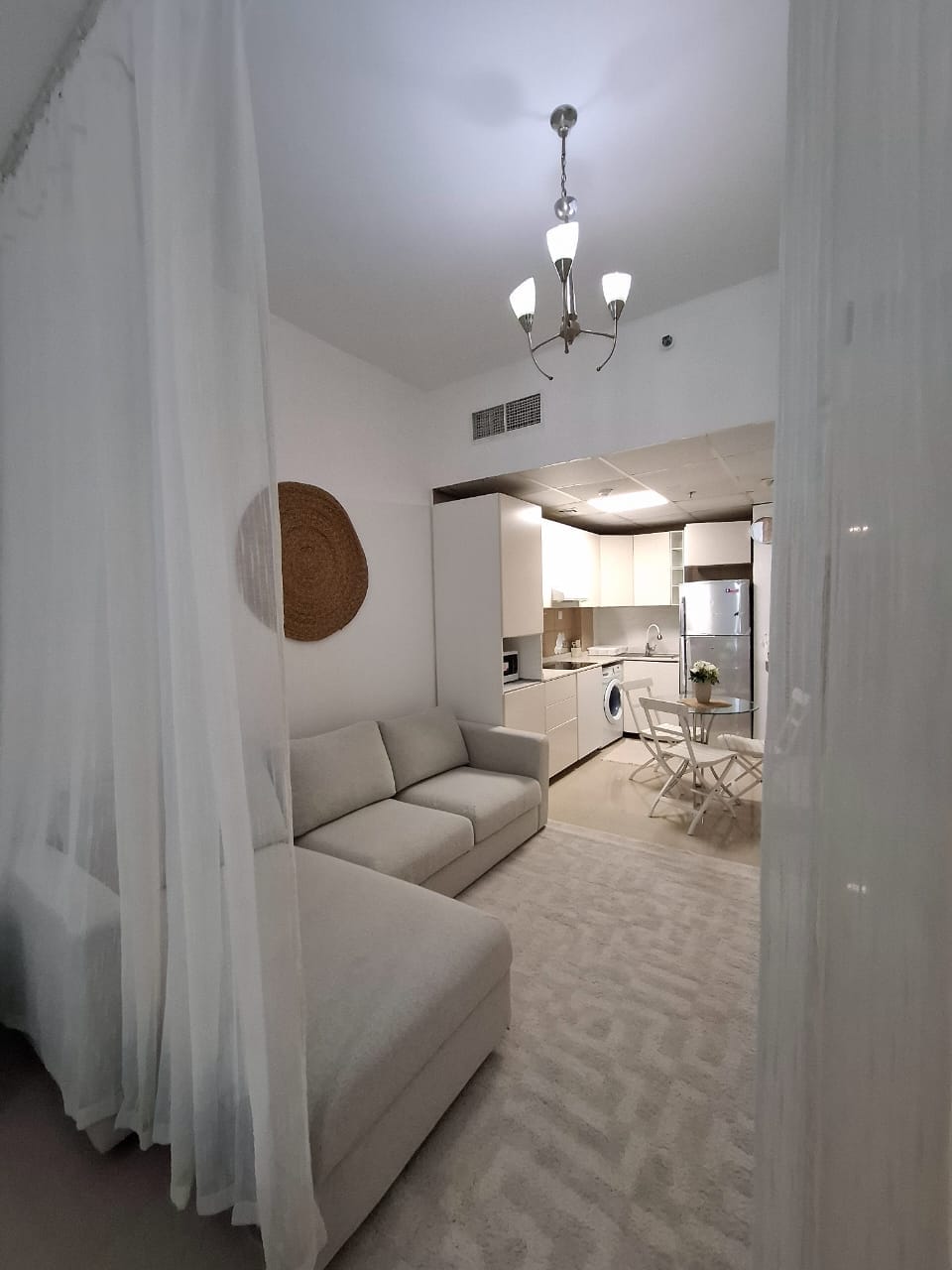 A modern living area is visible, featuring a light grey sofa and a circular decorative wall piece. The kitchen, equipped with white cabinetry and stainless-steel appliances, is adjacent to a compact dining set, all set against a neutral backdrop.