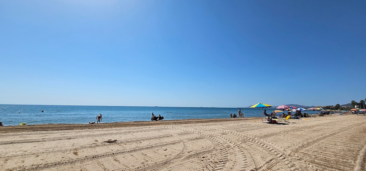 A sandy beach stretches under a clear blue sky, with gentle waves lapping at the shore. Sun umbrellas and beachgoers can be seen enjoying the expansive shoreline, while a few people are gathered in the water. Patterns of tire tracks lead across the beach sand.