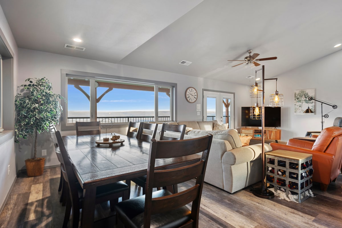 A spacious living area is presented with a large dining table and comfortable seating arrangements. Large windows offer ample natural light and views of the outdoors. Decorative elements include a potted plant and a wall clock, creating a welcoming environment.