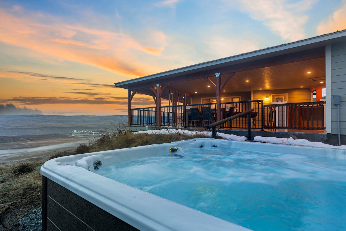 An outdoor hot tub, designed for eight people, is positioned at the edge of a deck. A sunset sky with orange and pink hues is visible in the background, complementing the scenic views of the landscape. The covered patio area features a dining set in the distance.