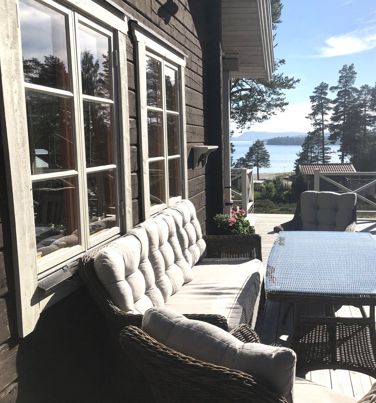 A patio area is visible, featuring a comfortable seating arrangement with a cushioned sofa and chairs. A dining table is set nearby. The view beyond showcases a scenic landscape of tall trees and water, with sunlight illuminating the space.