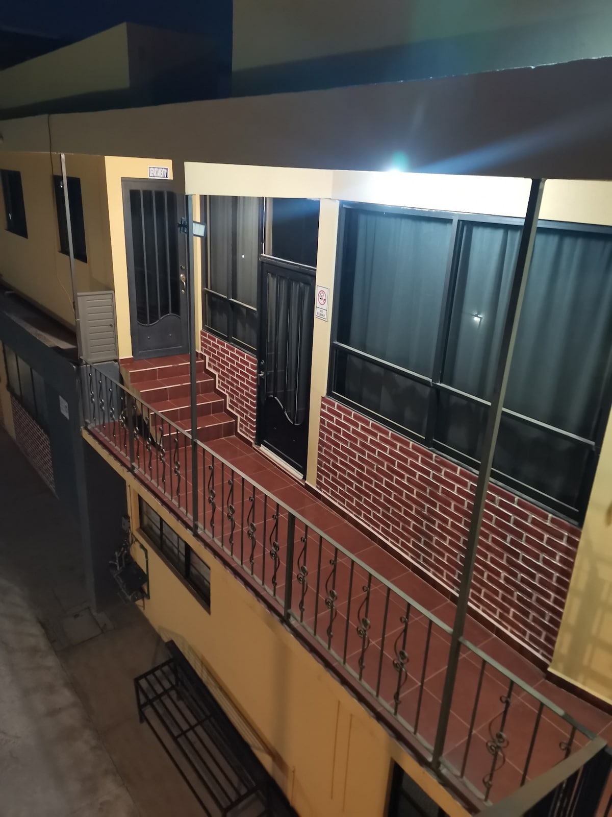 A well-lit exterior view of the second floor showcases a balcony with metal railing and a red brick facade. The entrance door is framed by large windows, allowing natural light into the area. A staircase leads down to the lower level, enhancing accessibility.
