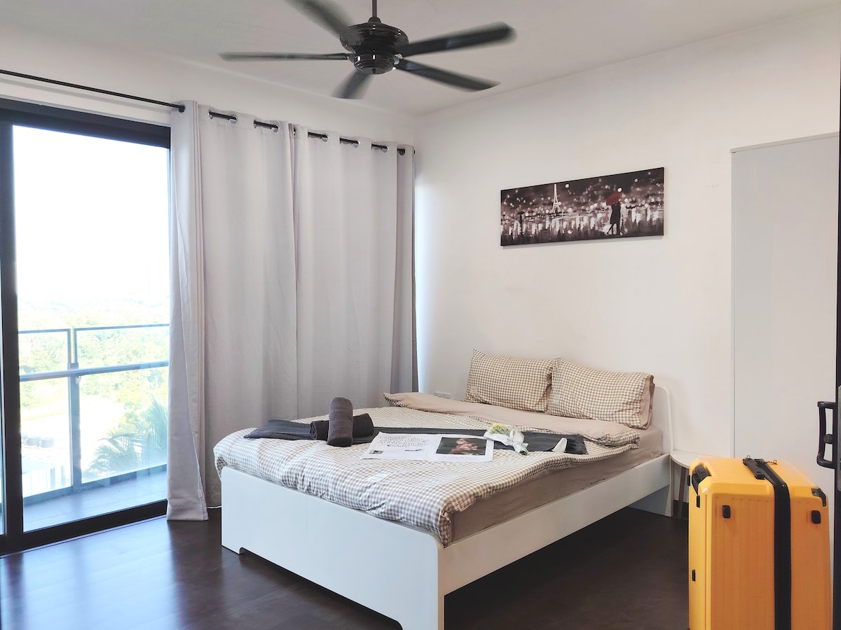 A bright bedroom is featured, showcasing a spacious bed layered with neutral-toned bedding. Light curtains frame a large window, offering a view. A yellow suitcase is positioned beside the bed, and towels are neatly arranged on the bedding.