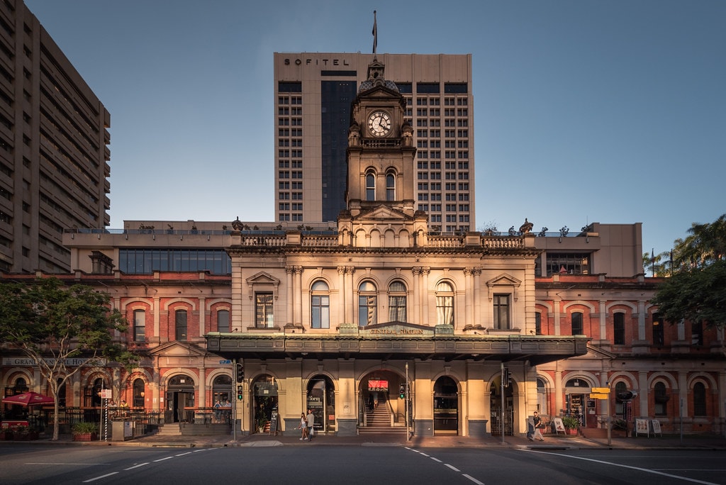 The historic building features classic architectural elements, with ornate detailing around the windows and a prominent clock tower at its peak. The façade is illuminated, highlighting the rich tones of the bricks and the arched entrance. Modern buildings are visible in the background.