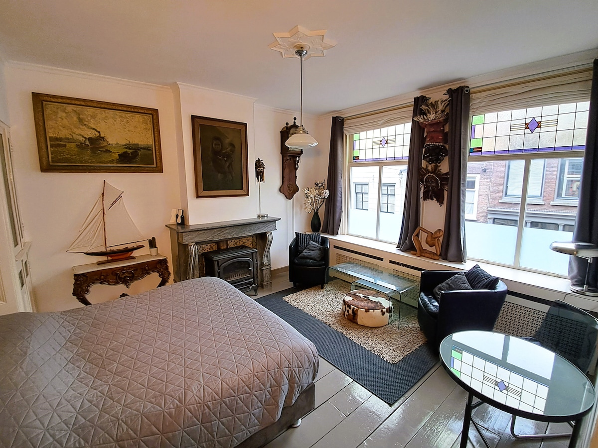 A stylish room features a spacious bed covered with a textured gray blanket. Two comfortable chairs are positioned beside a glass coffee table, with a decorative sailboat and artwork accentuating the walls. Stained glass above the window invites soft natural light.