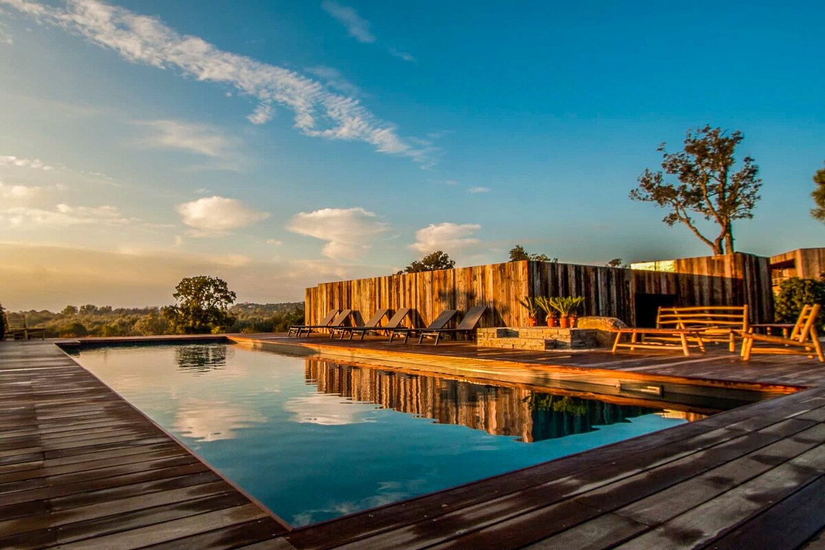 A spacious outdoor pool reflects the blue sky and surrounding landscape. Wooden decking encircles the pool, with several lounge chairs positioned for relaxation. Natural greenery and wooden structures are visible in the backdrop, contributing to a serene outdoor environment.