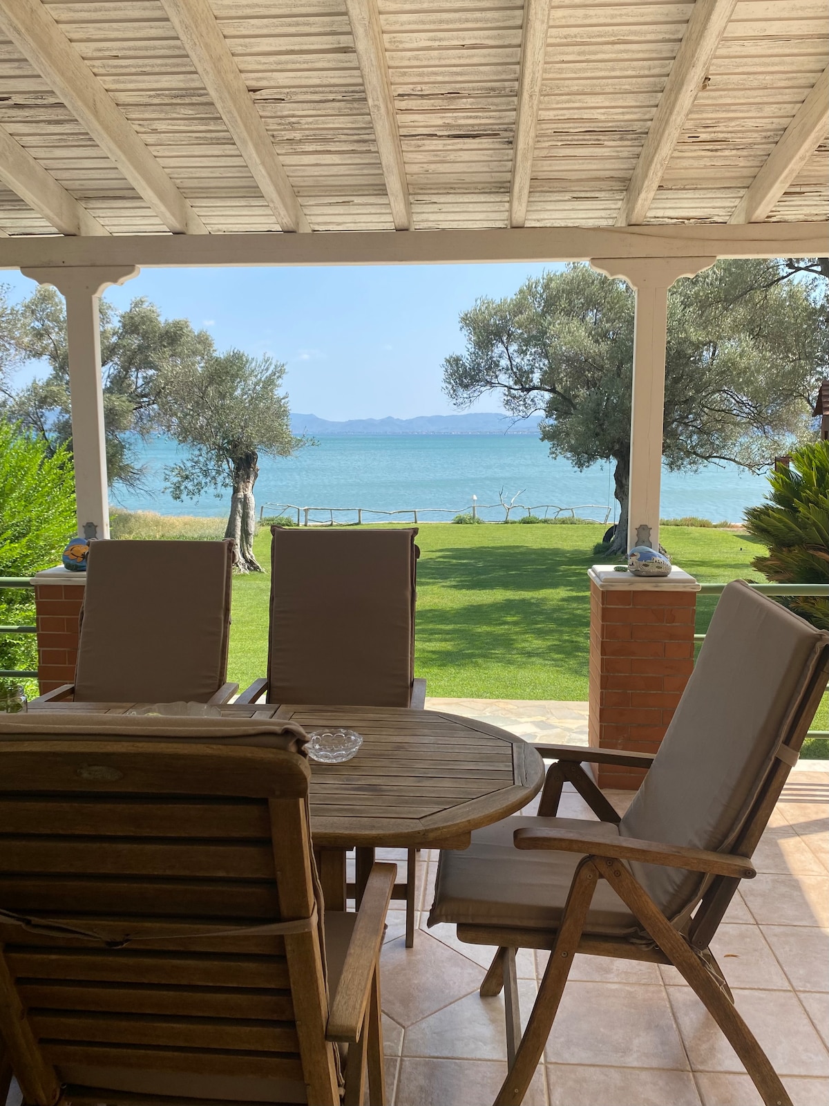 A shaded patio area is visible, featuring a circular wooden table surrounded by four comfortable chairs. Beyond the patio, a lush green lawn leads to the serene blue waters of the sea, framed by olive trees and distant hills.