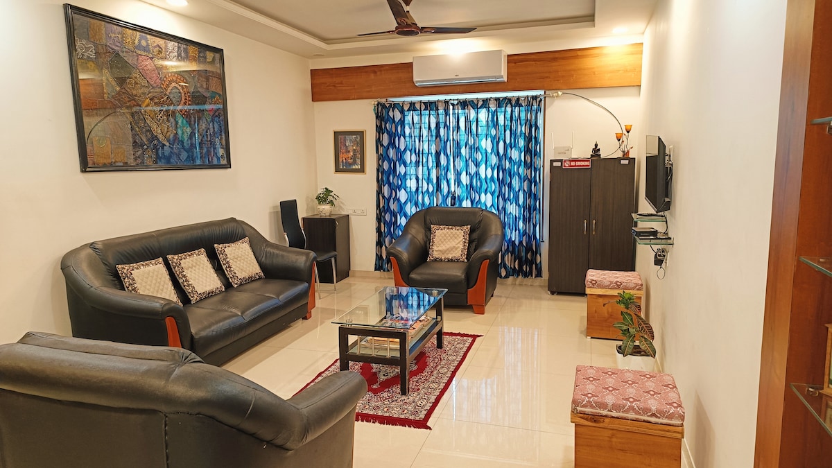 A welcoming living area features two black leather sofas and a matching armchair, arranged around a glass coffee table. Natural light filters through a patterned blue curtain. A decorative wall hanging adds color, and a wooden cabinet provides storage space.