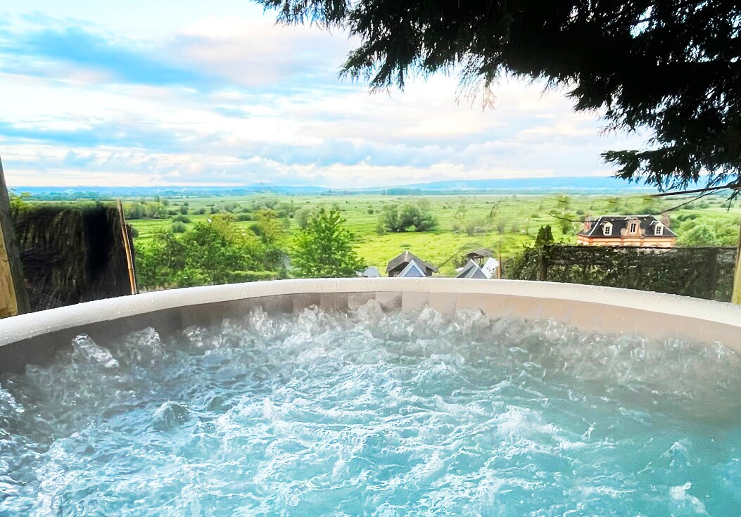 A bubbly outdoor jacuzzi is positioned on a wooden platform, offering a scenic view of expansive green fields and distant hills. The water glistens under the soft light, creating a tranquil setting for relaxation in nature.